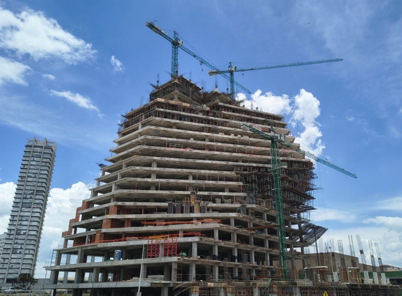 A concrete tower under construction against a blue sky, twelve floor plates cast, scaffolding and formwork on the upper levels, and two tower cranes standing over it.